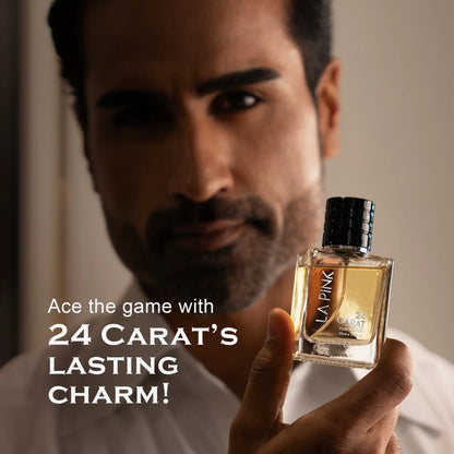 A man holds a bottle of LA PINK 24 CARAT fragrance, showcasing the clear glass bottle filled with amber-colored liquid and a textured black cap. The background is a softly blurred warm-toned interior, with promotional text on the left reading &