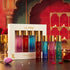 LA PINK Unisex Parfum Gift Set featuring a white box containing five 10ml travel perfume bottles with gold caps, displayed alongside five matching bottles on a dark wooden table with warm, decorative background elements.


