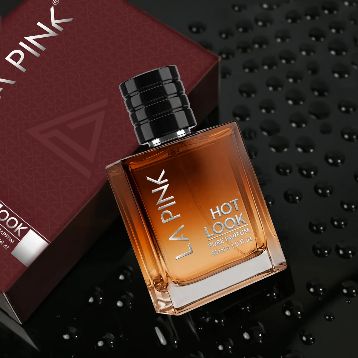 A bottle of La Pink Hot Look Pure Parfum with amber liquid and a black cap, shown next to its dark red box on a glossy black background with water droplets.

